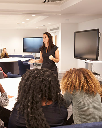 A lecturer (standing) instructs a group of university students (sitting)