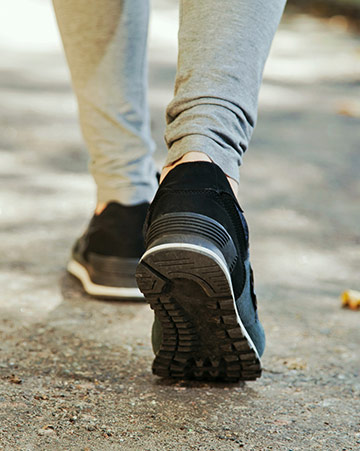 Person walking outdoors on a paved path, wearing grey joggers and black trainers, viewed from behind with one foot lifted mid-step.