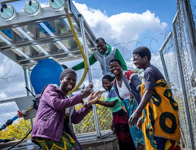 Group of five people in Malawi gathered outdoors beside a metal-framed water system with a large blue tank and yellow pipe.