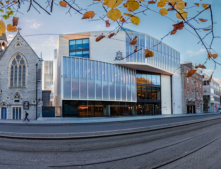 Contemporary glass-fronted RCSI building beside a historic stone church, viewed across tram tracks with autumn leaves framing the scene.