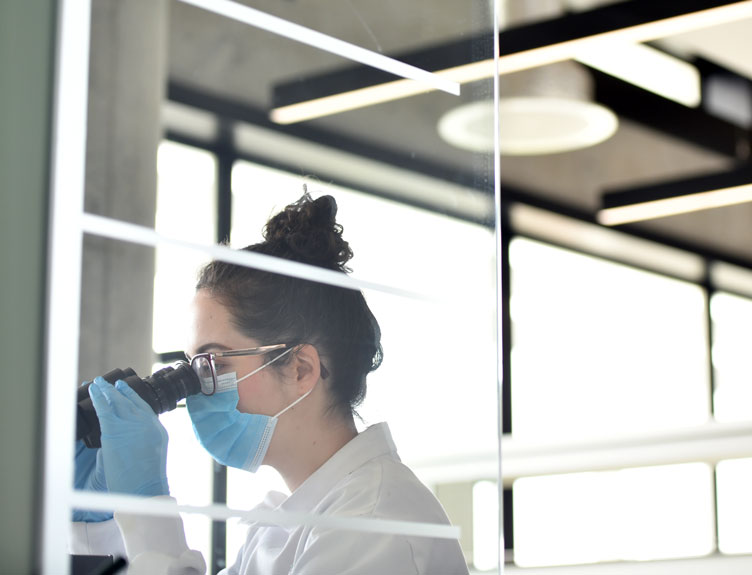 A laboratory worker wearing a face mask and gloves looks through a microscope in a bright, modern research facility with large windows.