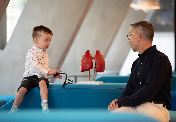 Child and adult seated opposite each other with a model of human lungs between them, discussing respiratory health in an educational or healthcare setting.