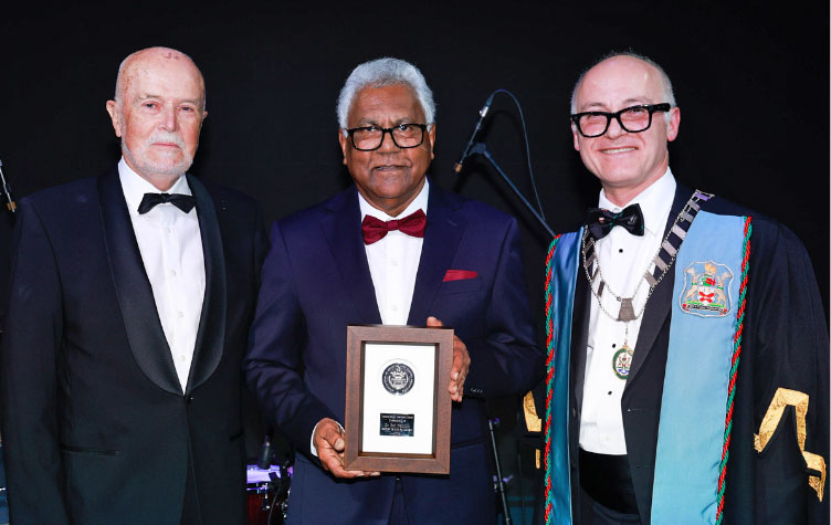 Three people in formal evening attire stand on a stage in front of a dark backdrop. The person in the centre holds a framed award plaque.