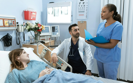 Patient resting in a hospital bed while a healthcare professional reviews information on a clipboard and another person sits beside the bed in a clinical care environment.