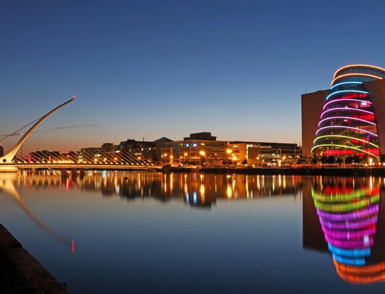 Night view of Dublin’s River Liffey with the Samuel Beckett Bridge on the left and the Convention Centre Dublin on the right, illuminated with multi-coloured lights reflected in calm water.