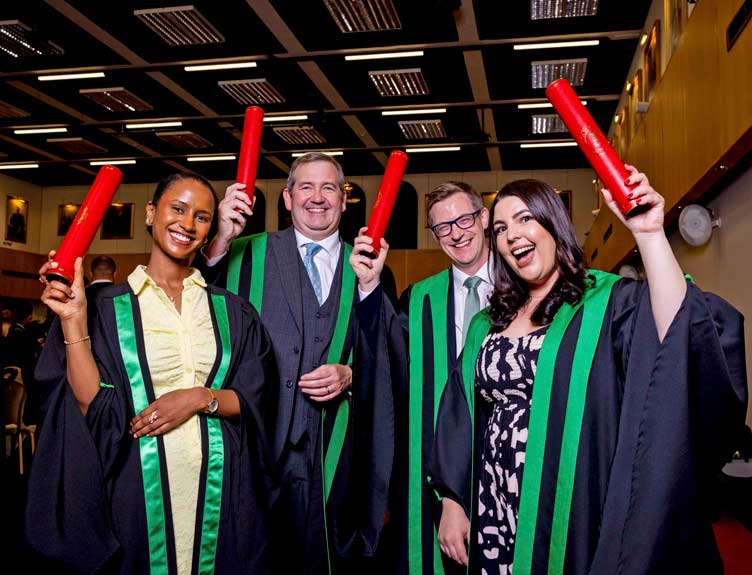 Group of four people standing indoors at a graduation or award ceremony, wearing black robes with green trim and holding red diploma tubes. 