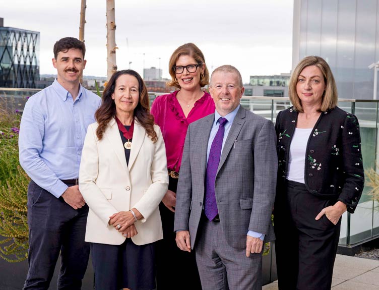 A group of five professionals standing together on an outdoor terrace in a corporate or academic setting, with modern buildings visible in the background.