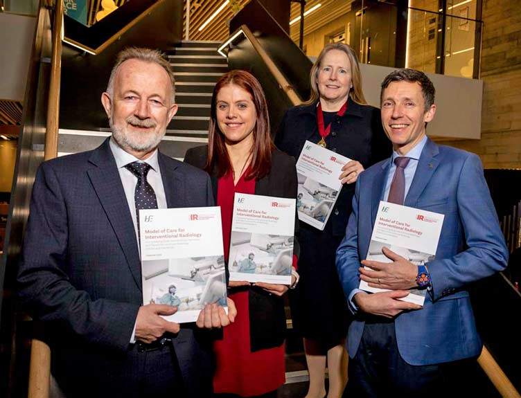 Four people hold printed copies of the Model of Care for Interventional Radiology in front of staircase