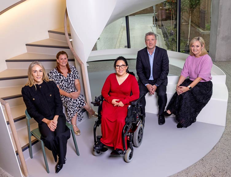 Five people seated on a curved staircase in a modern building, including one wheelchair user in a red outfit at the centre, posing for a group portrait.