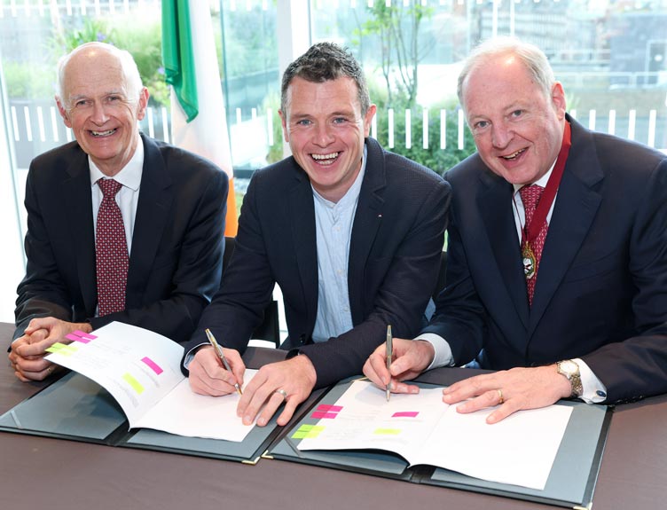 Three individuals dressed in business suits seated at a table indoors, signing documents placed inside open folders.