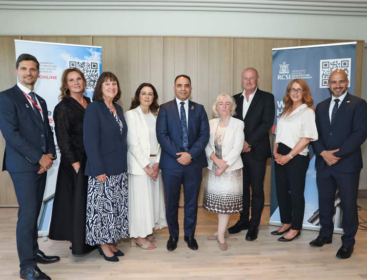 Nine people in business attire pose for a group photo between University of Medicine and Health Sciences and RCSI Graduate School of Healthcare Management display banners at an indoor event venue.