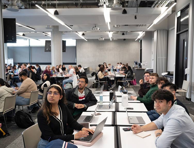 Students working on laptops in a modern classroom, seated at group tables during a collaborative workshop or study session.