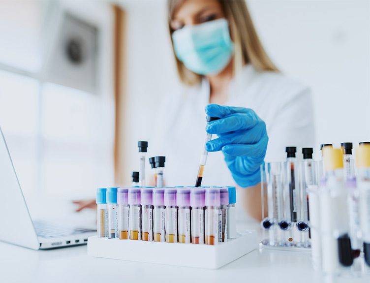 A scientific researcher wearing blue gloves and a mask holds up a blood sample