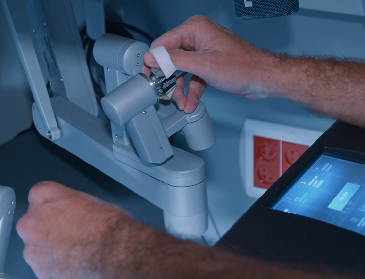 Close-up of a hand operating the control mechanism of a robotic surgical system, with a touchscreen control panel visible beside the equipment.