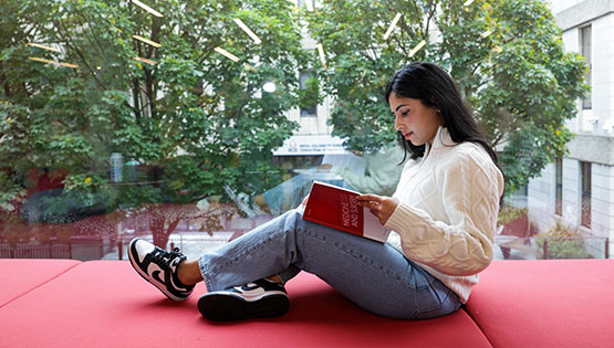 Student reading a book while seated in an RCSI campus study area beside a large window overlooking trees and university buildings.