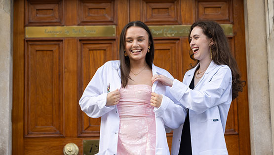 Two students wearing white coats standing outside an RCSI university building, adjusting the coats after a white coat ceremony.