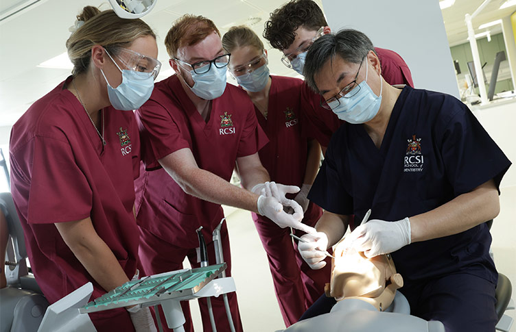 A group of RCSI Dentistry students dressed in maroon scrubs are shown how to examine teeth on a patient simulator by a lecturer