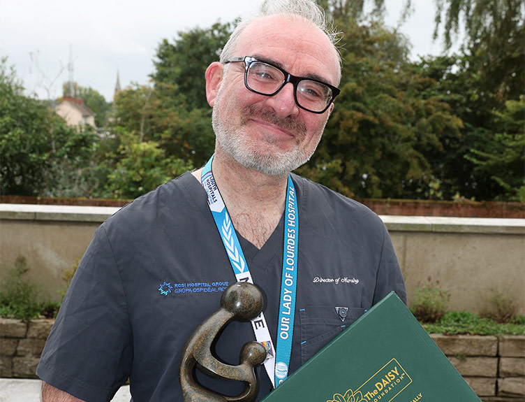 A healthcare professional in scubs holding a DAISY Award certificate and award statue.
