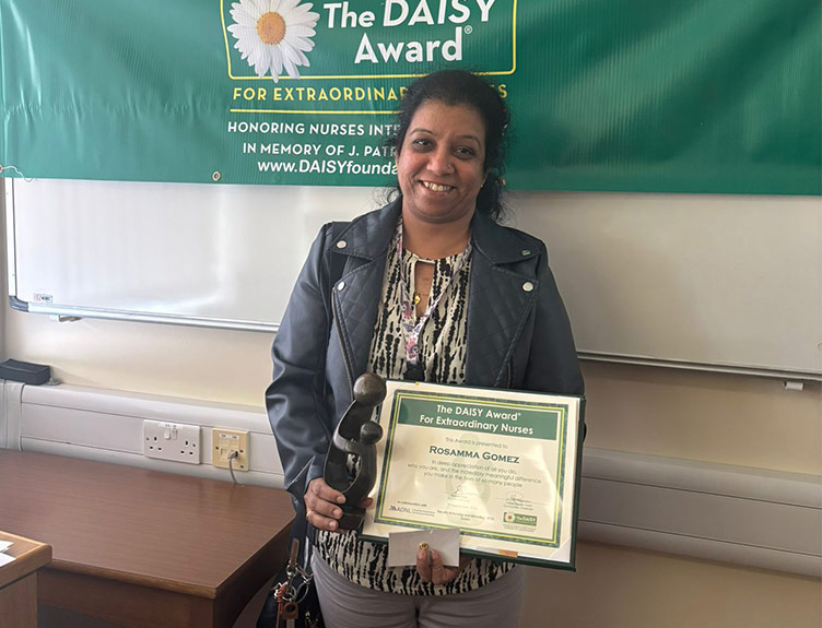 A person, standing in front of a DAISY banner, holds a DAISY Award certificate and award statue.