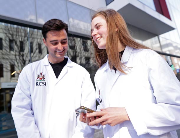 Two students in white coats converse outside RCSI building