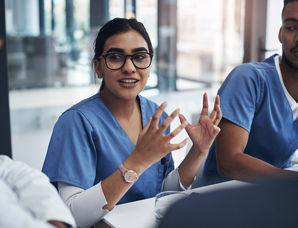 A young doctor wearing scrubs gesticulates during a meeting with colleagues.