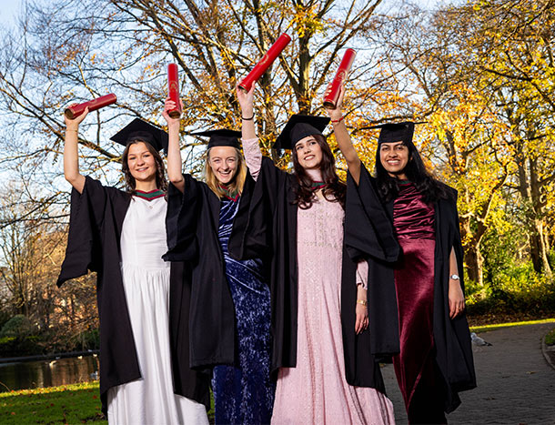 Graduates in academic gowns and mortarboards holding ceremonial degree scrolls aloft while posing outdoors after a conferring ceremony.