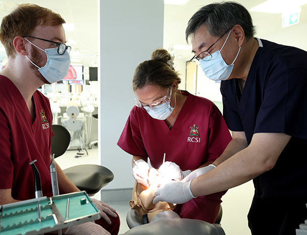 Two RCSI Dentistry students learn from a tutor in a similated environment. All are wearing scrubs.