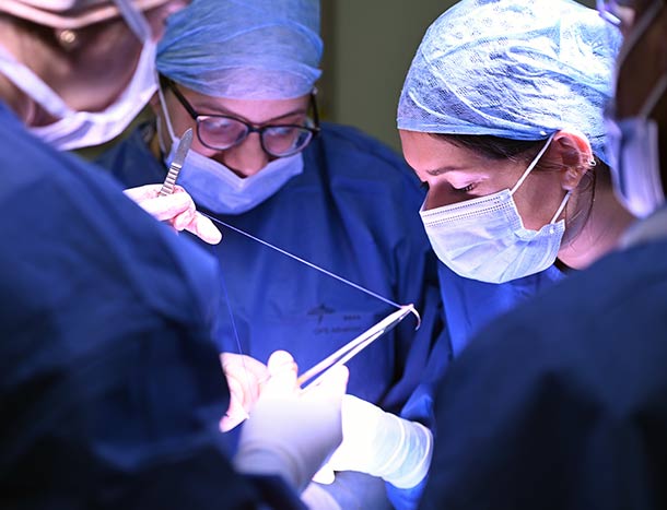A surgeon holds a suture while being directed by another surgeon