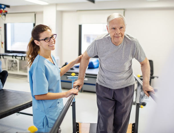 A nurse assists an elderly gentleman supporting himself on a walking rail.