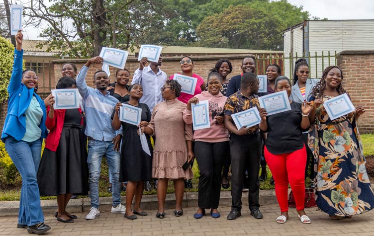 Group of Akazi certificate recipients posing outdoors and holding up their certificates during a training or graduation celebration.