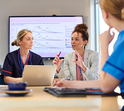 Three female managers and healthcare professionals at a table around a laptop, with a big-screen presentation in the background.