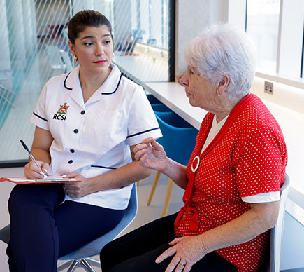A healthcare professional in an RCSI uniform speaks with an older patient during a consultation, reviewing information together in a bright clinical setting.