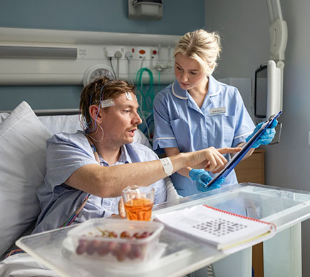 A nurse with a clipboard assists a patient who is sitting up in bed.
