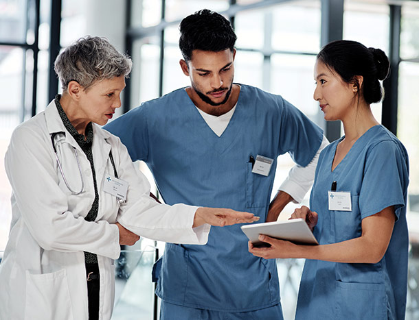 Three healthcare professionals in a hospital corridor reviewing patient information together, with one pointing to a document while another holds a tablet.