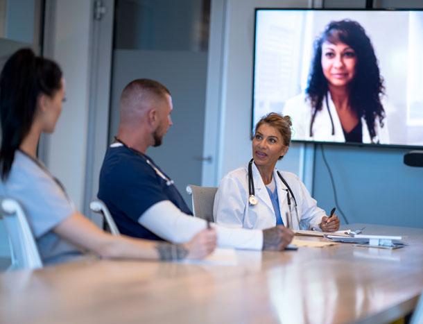 Healthcare professionals seated around a conference table participating in a video meeting, with a remote participant displayed on a screen at the front of the room.