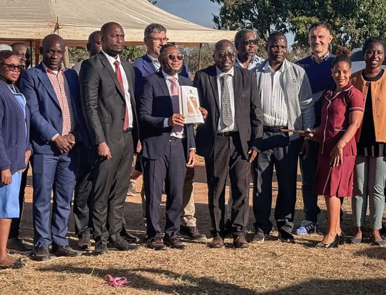 Group photo of people in business, healthcare, and work attire standing outdoors in front of a canopy tent, with one person holding a framed certificate during an official presentation or recognition event.