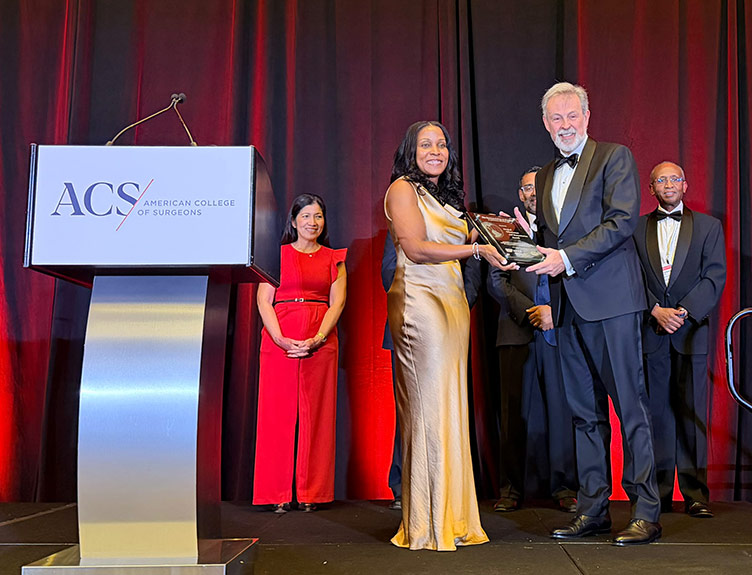Individual receiving a recognition award on stage at an American College of Surgeons event, with other attendees standing nearby and an ACS-branded podium visible in the foreground.